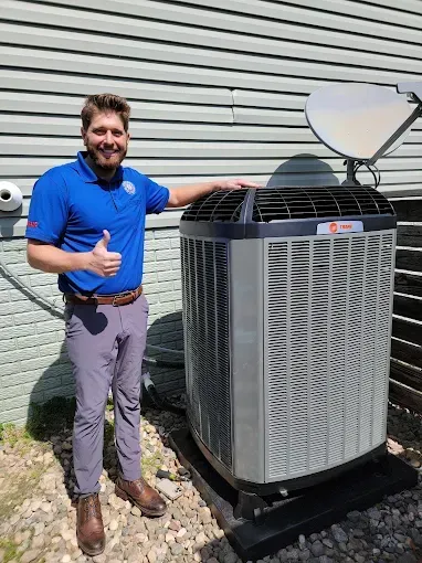 Man in blue shirt, thumbs up, standing next to a Trane air conditioning unit on a gravel base.