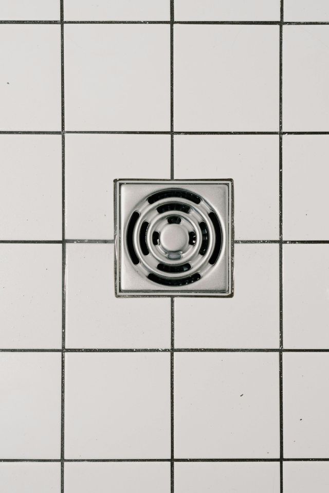 Square chrome shower drain centered on white tiled floor.