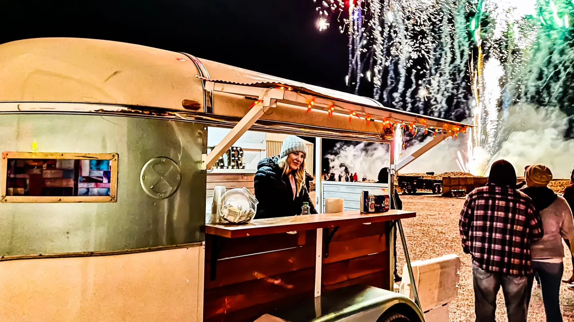 A man is standing in front of a food truck with fireworks in the background.