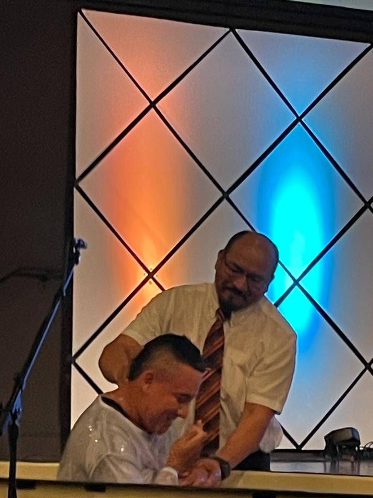 Man being baptized in a church. A man in a white shirt assists, while colorful lights shine behind.
