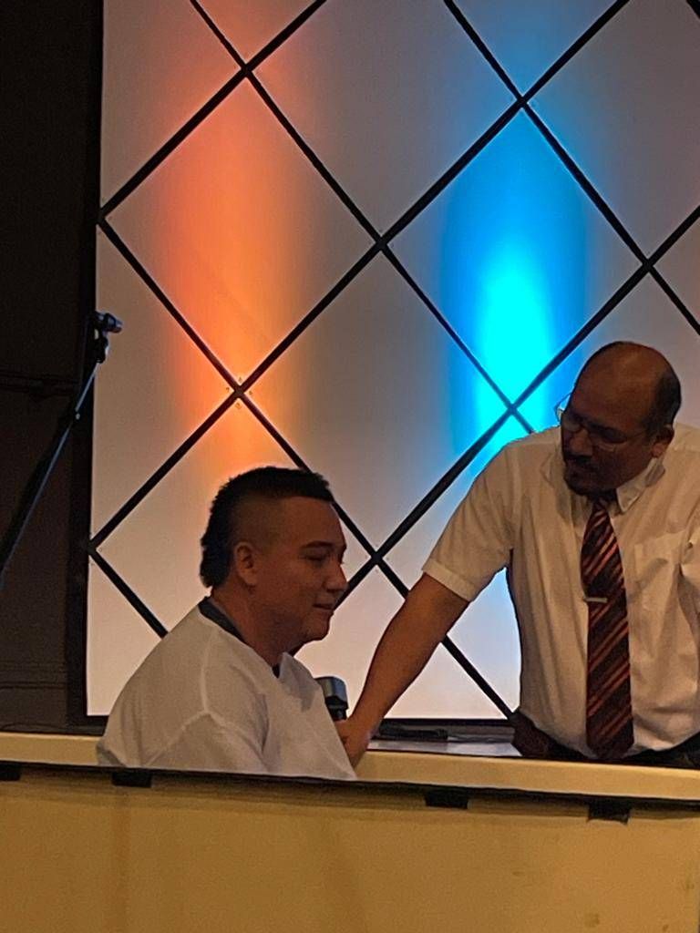 Man being baptized in a church. A man in a white shirt assists him near a tank, with orange and blue lights in the background.