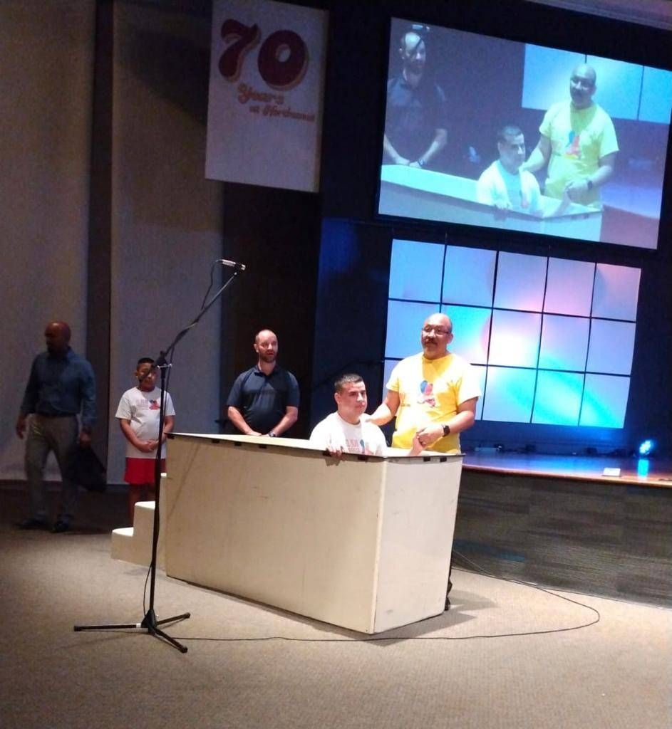 Baptism ceremony on a stage. Man in yellow shirt baptizes another in white tank top. Other men and a screen in background.
