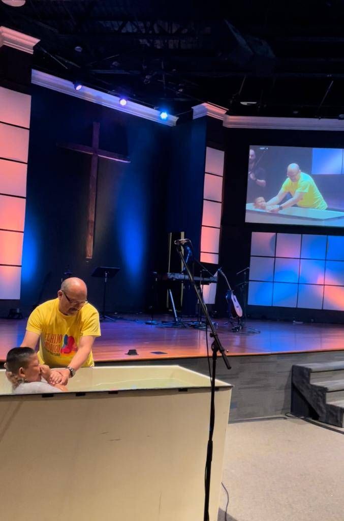 A person being baptized in a church. A man in yellow assisting in a baptismal. Stage is lit with blue and orange.