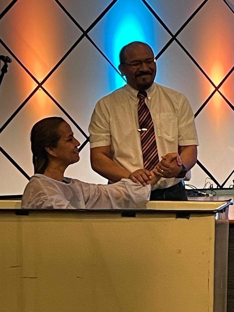 Person being baptized in a tub, held by a man. Background is lit with blue and orange.