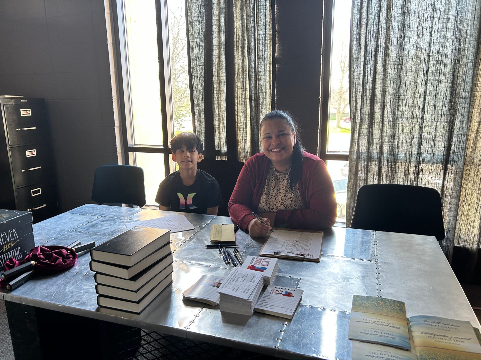 Two people sit behind a table. A stack of books, and cards are on the table. Sunlight streams through a window.
