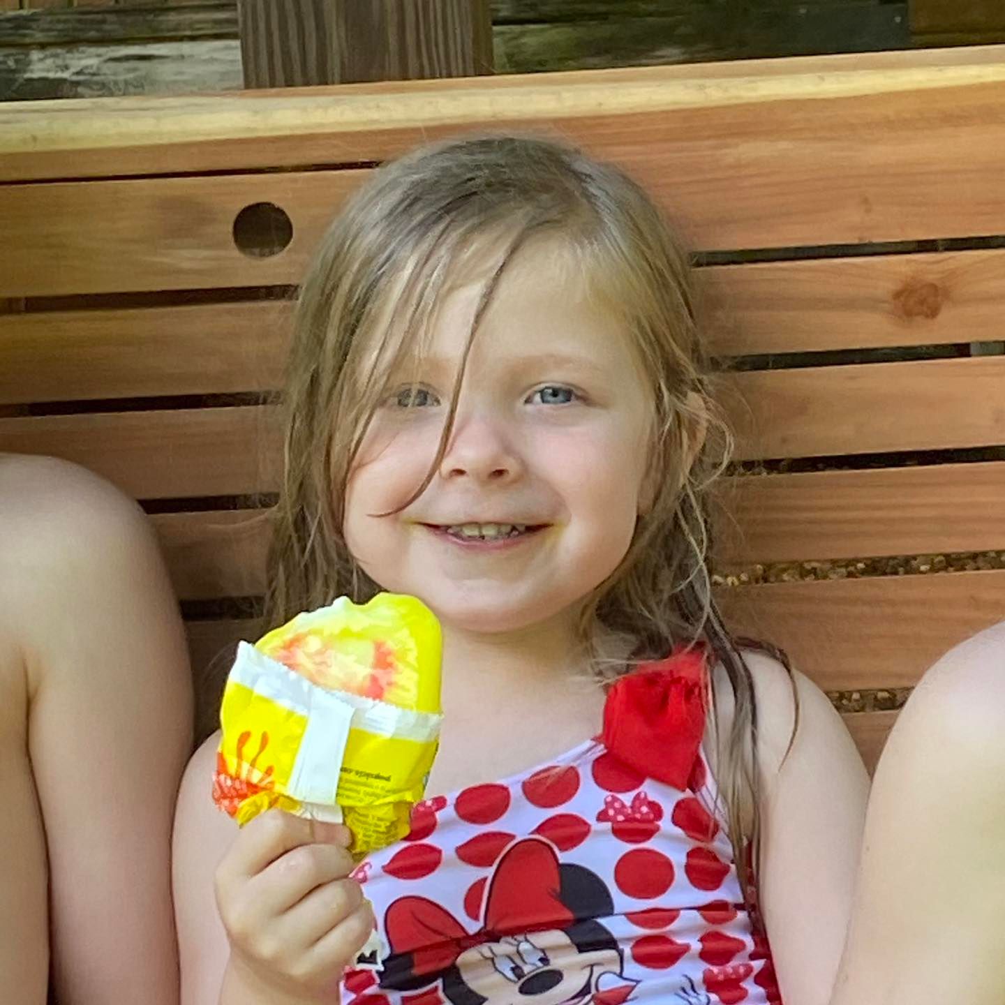 Young girl in a polka-dot swimsuit smiles, eating a wrapped ice pop on a wooden bench.