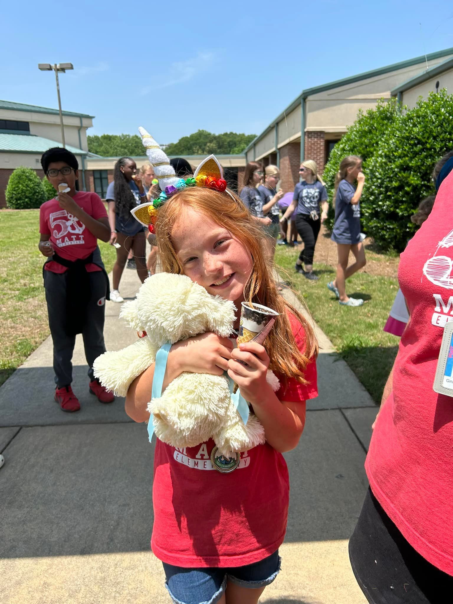 Girl with red hair wearing a unicorn headband hugs a teddy bear, smiling. Outside, holding ice cream.