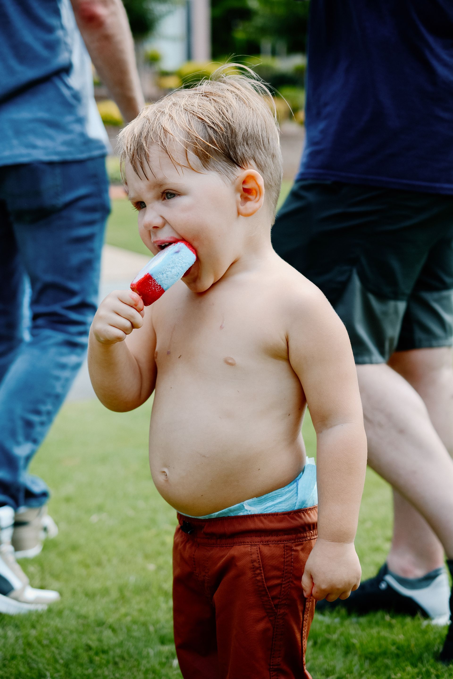 Bare-chested toddler eats a popsicle outdoors, wearing brown pants and a diaper.