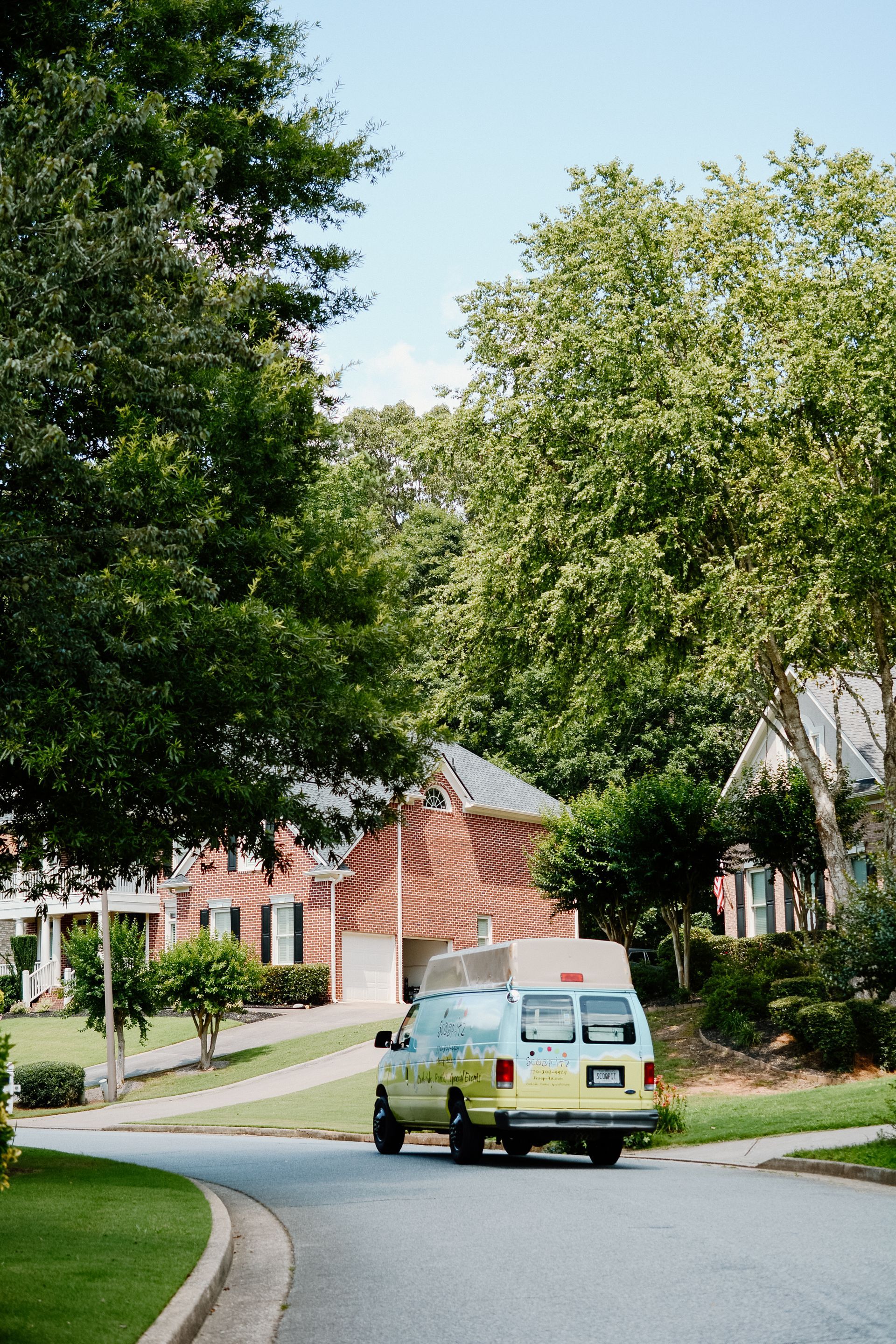 A yellow van carrying a box drives down a residential street lined with green trees and houses.