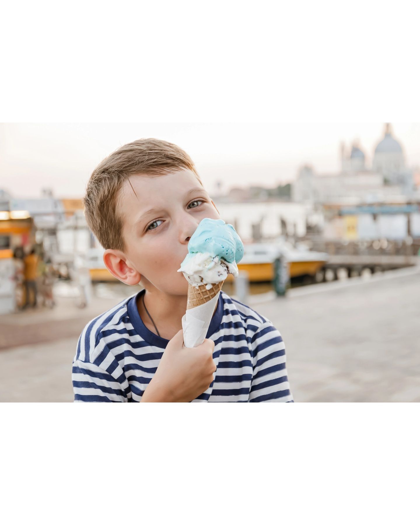 Boy eating ice cream cone with blue and white scoops near a waterfront.