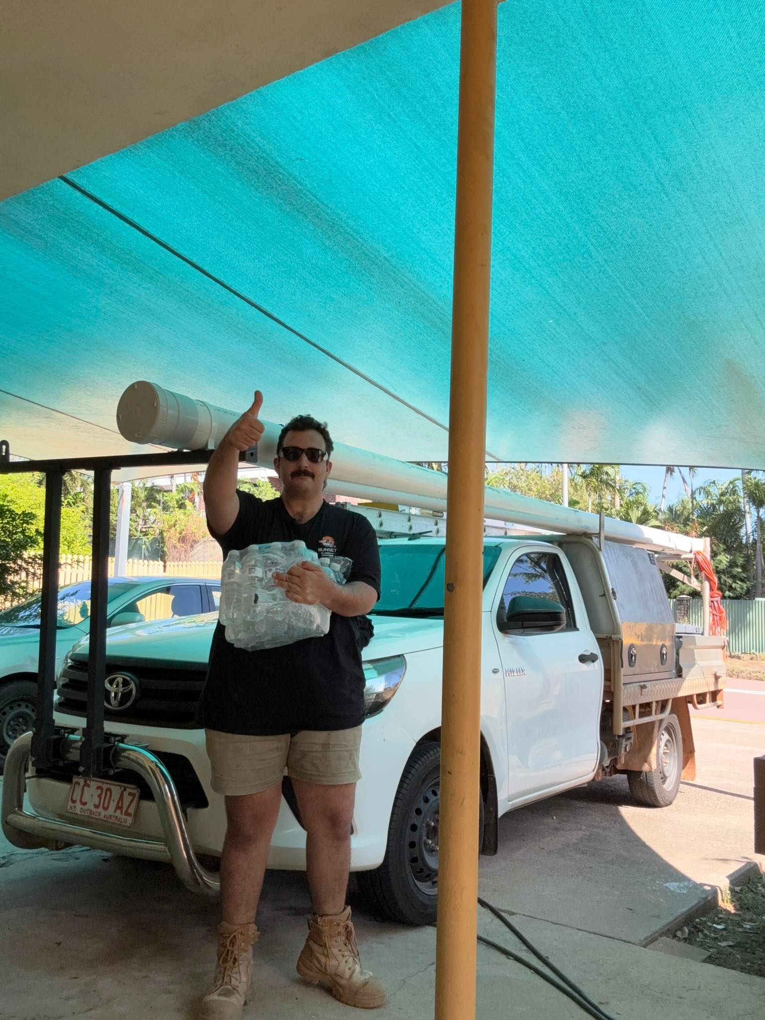 A man is standing in front of a white truck under a shade.