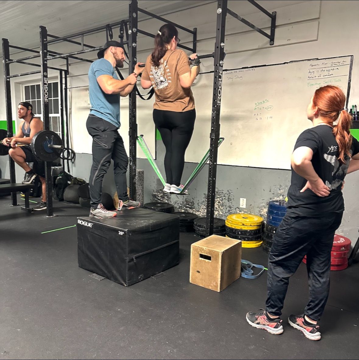 People on balance beams in a gym, one being coached while standing on a box.