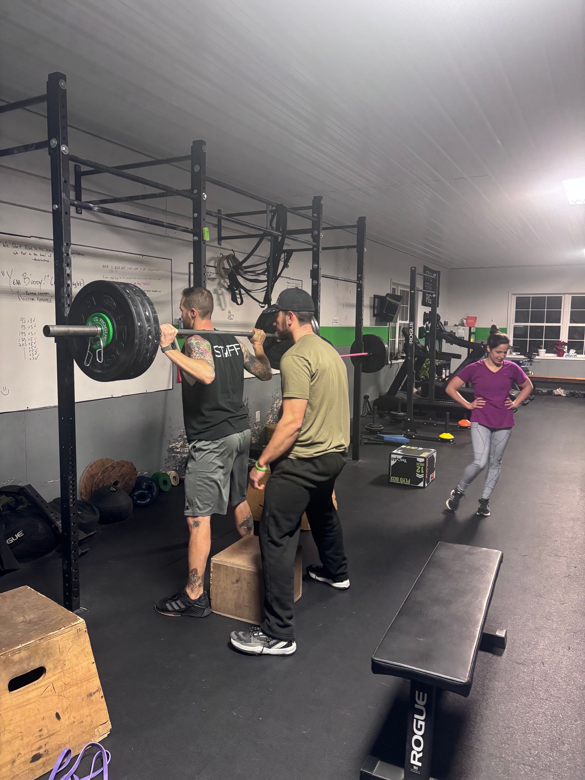 People lifting a barbell in a gym, with a coach nearby and another person watching in the background.