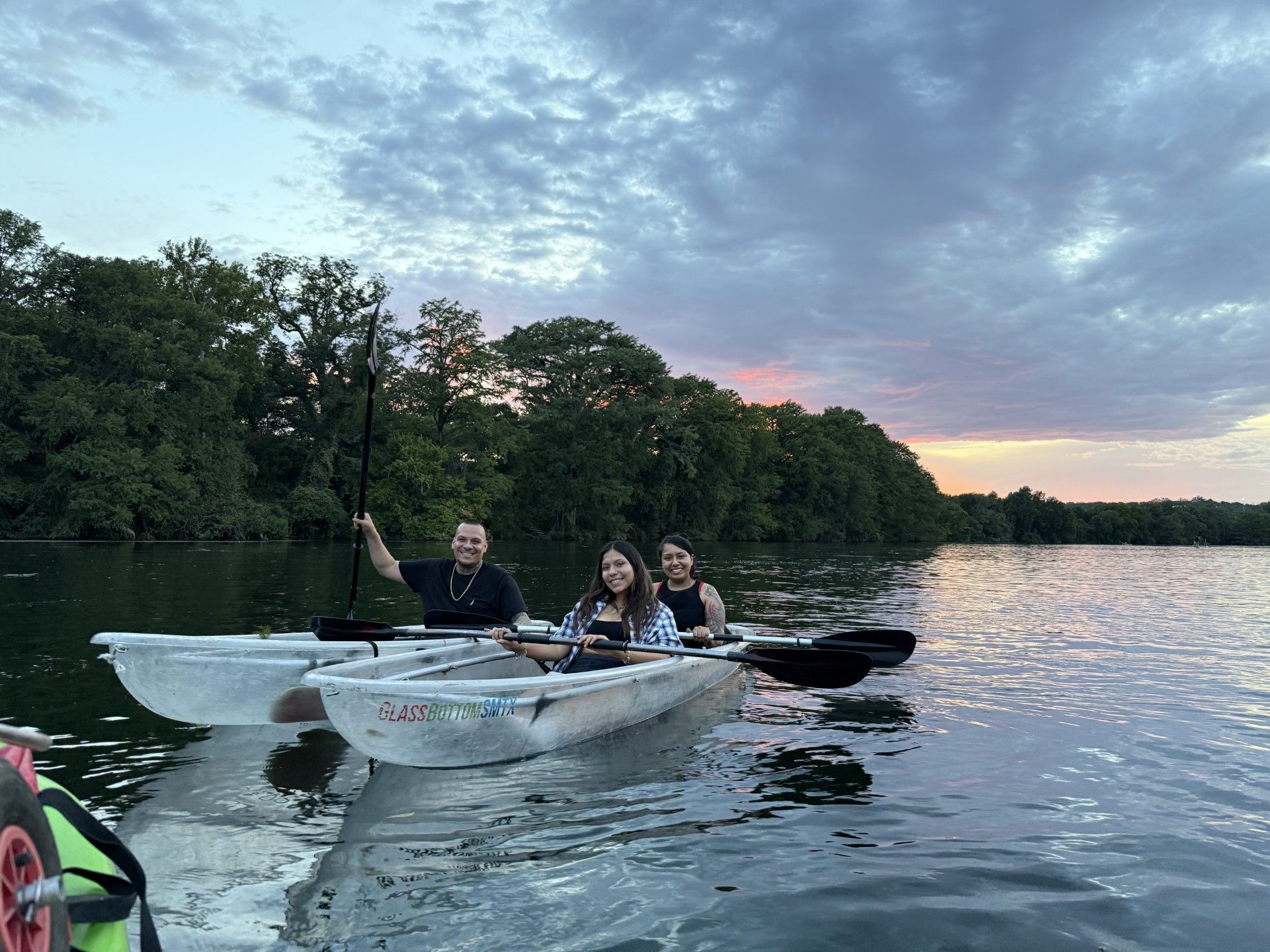 Three people kayaking on calm water at dusk, trees in background.