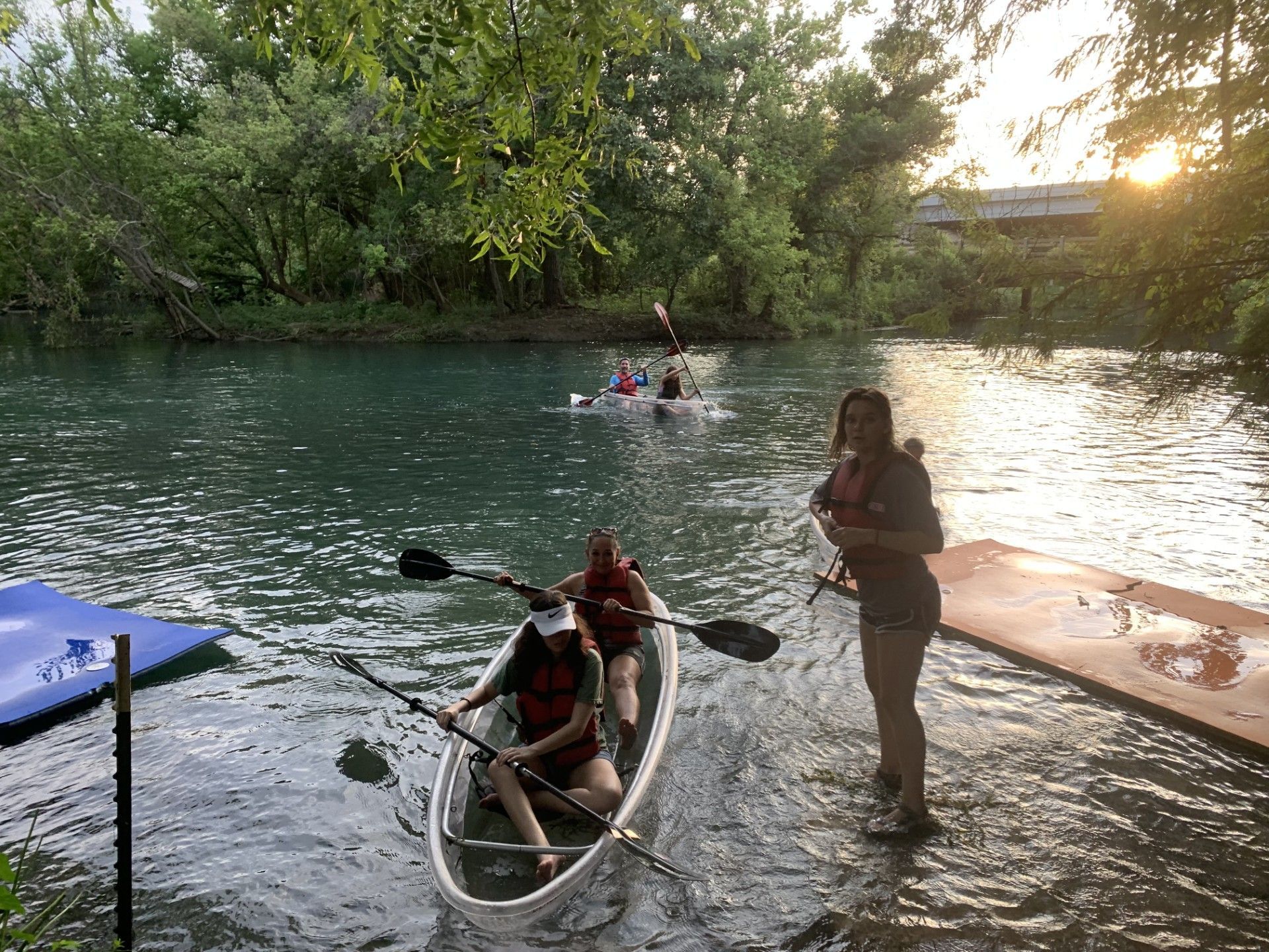 People kayaking on a river. One clear kayak, a dock and trees in the background, sunset.