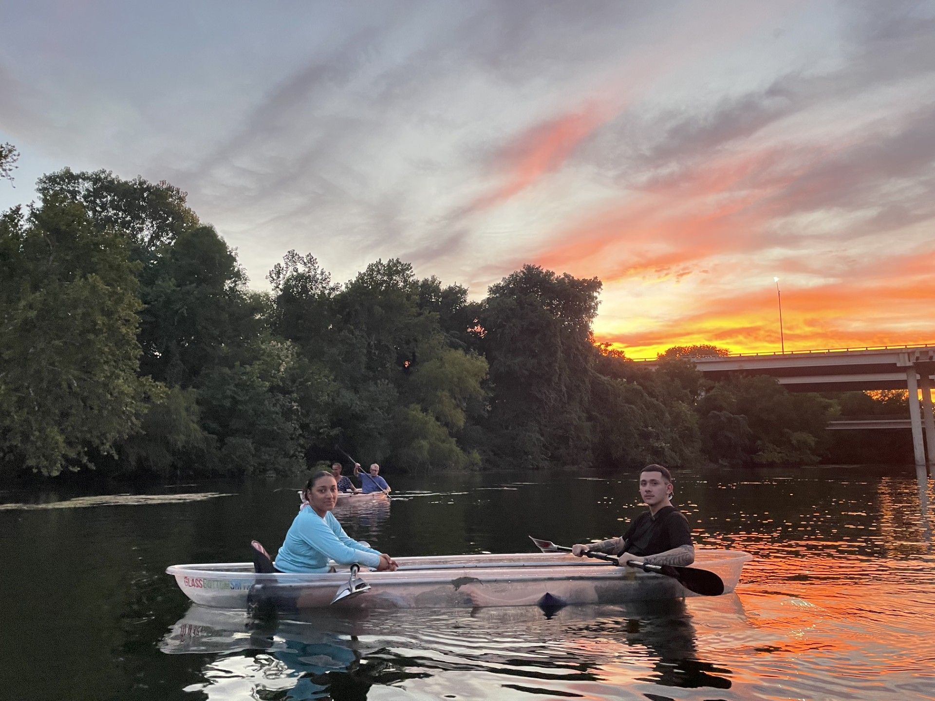 People in kayaks on a lake at sunset; orange and pink sky reflecting in the water; trees line the shore.