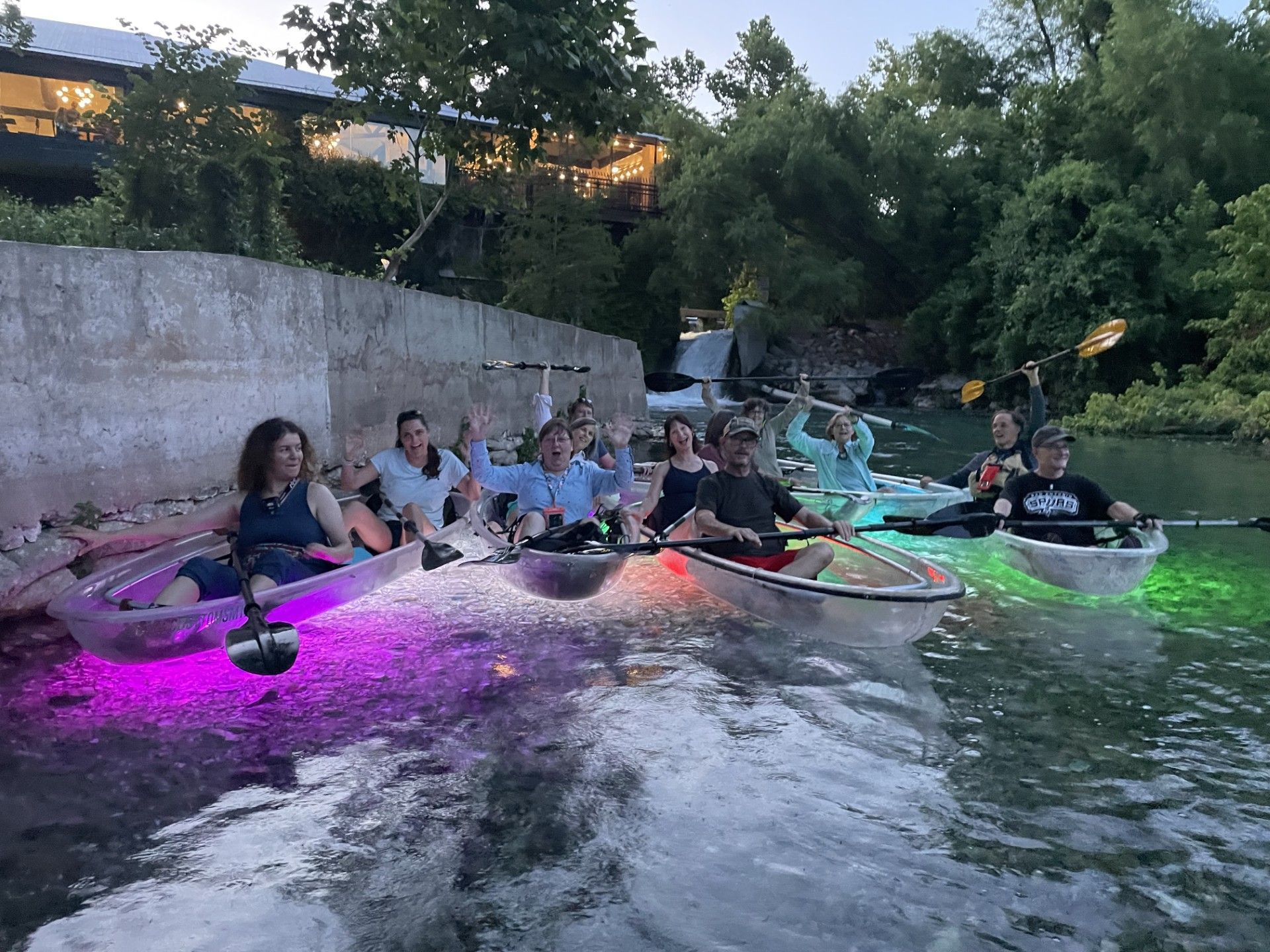 People in illuminated, clear kayaks on a waterway at dusk, waving toward the camera.