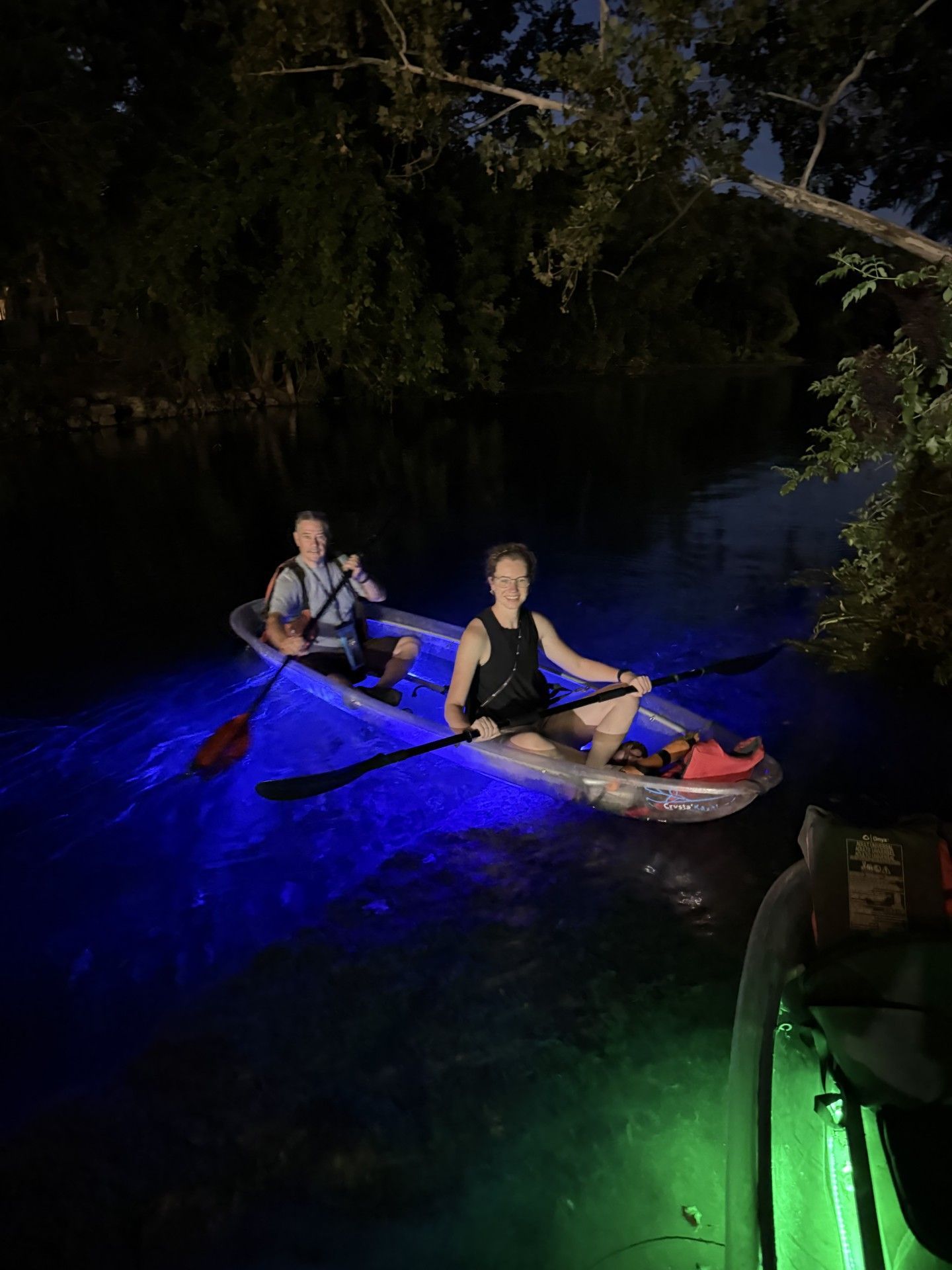 Two people in a clear kayak with blue lights on a dark waterway at night.