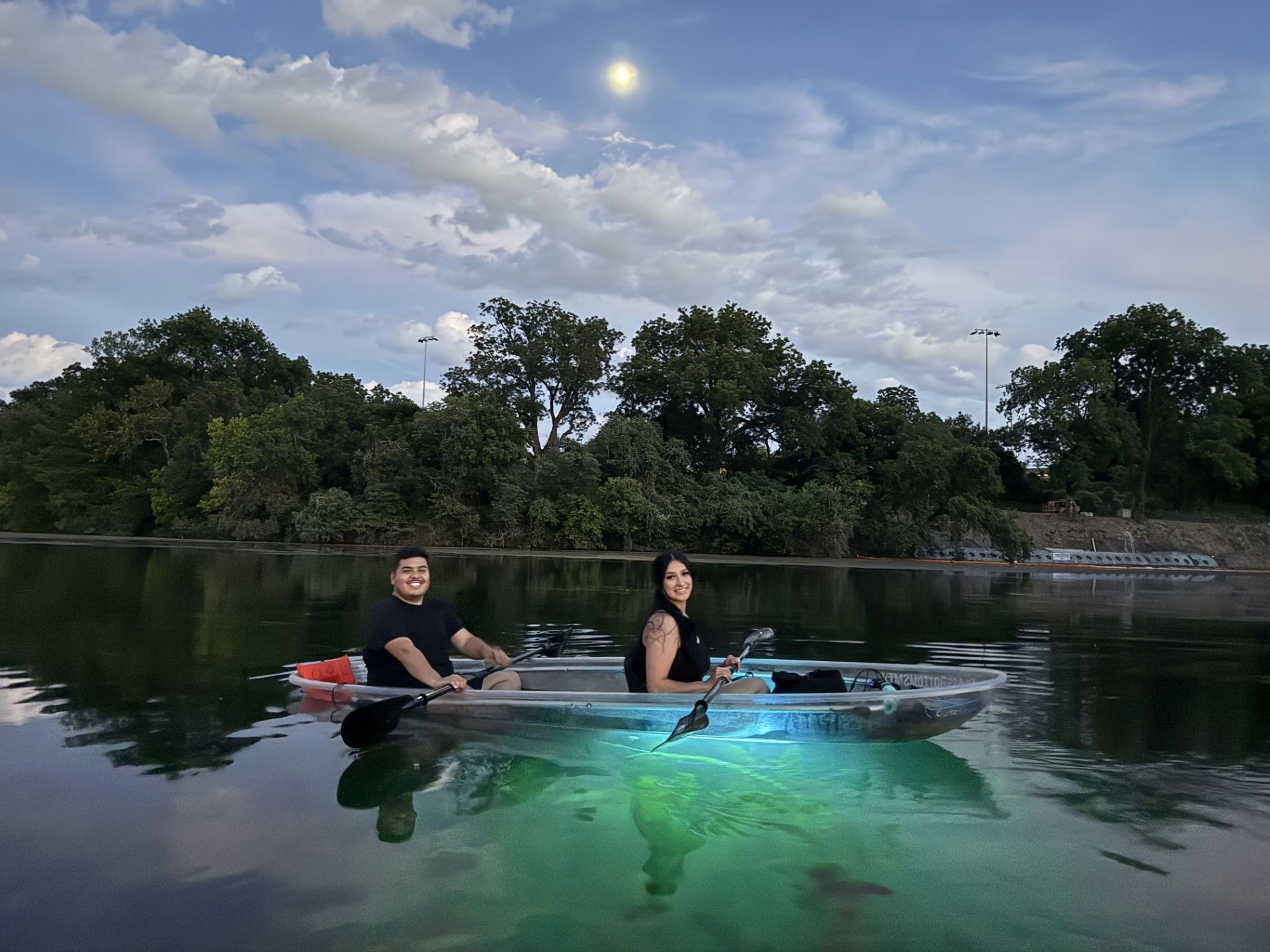 Two people kayaking in a clear kayak on a lake at dusk. Green lights illuminate below, with trees in the background.