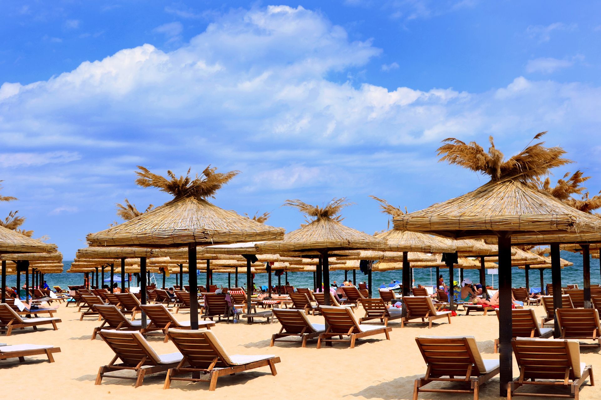 Beach with rows of straw umbrellas, lounge chairs, and people under a blue sky with clouds.