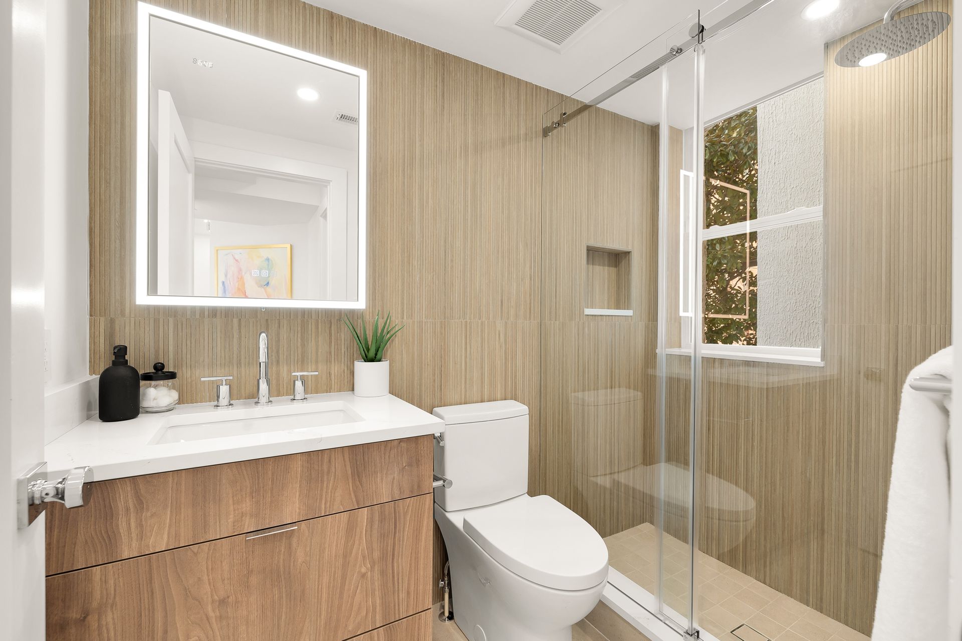Bathroom with wood-look paneling, lit mirror, glass shower, toilet, and a small window.