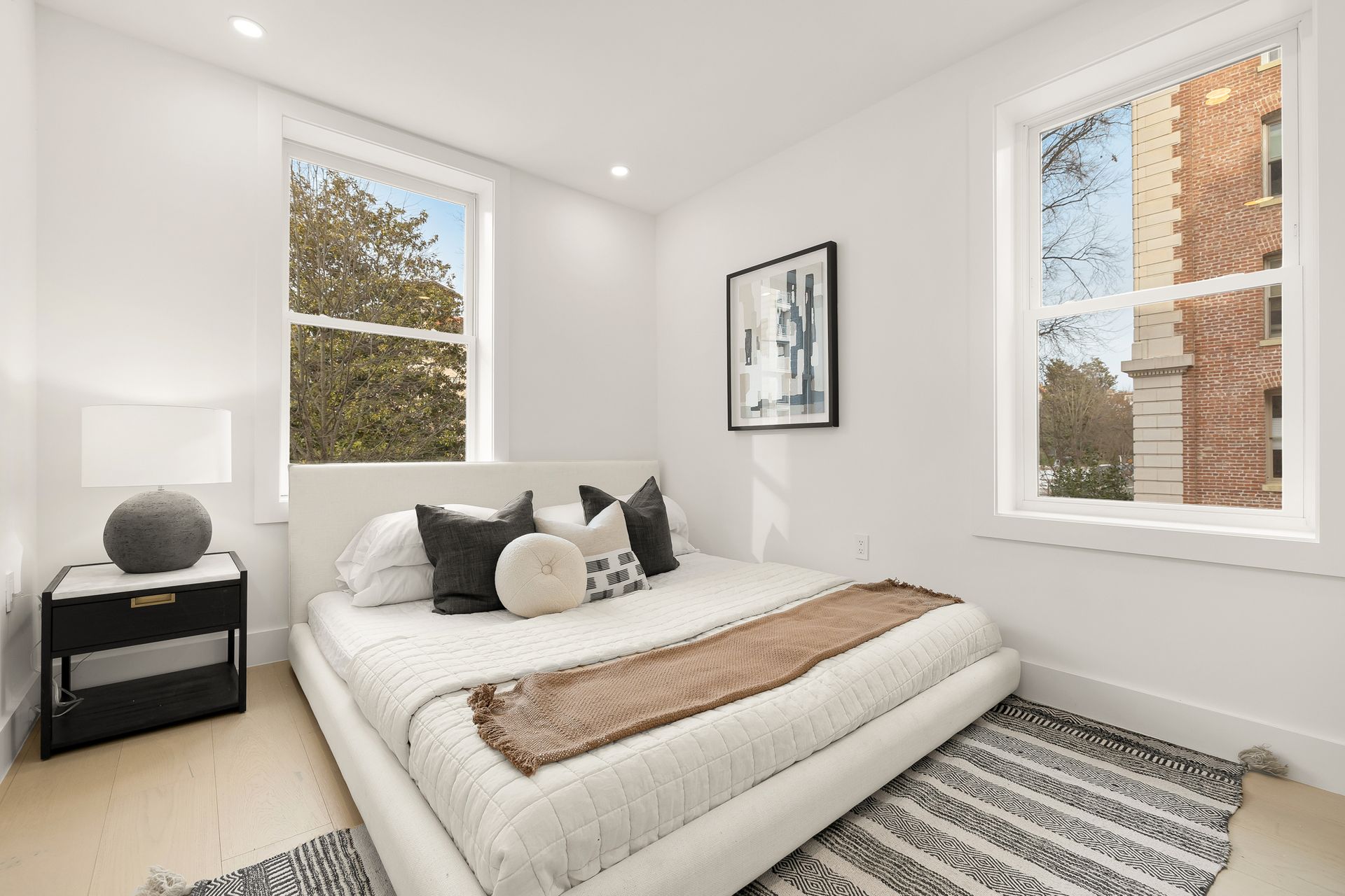 White bedroom with a bed, two windows, a nightstand, and a framed artwork.
