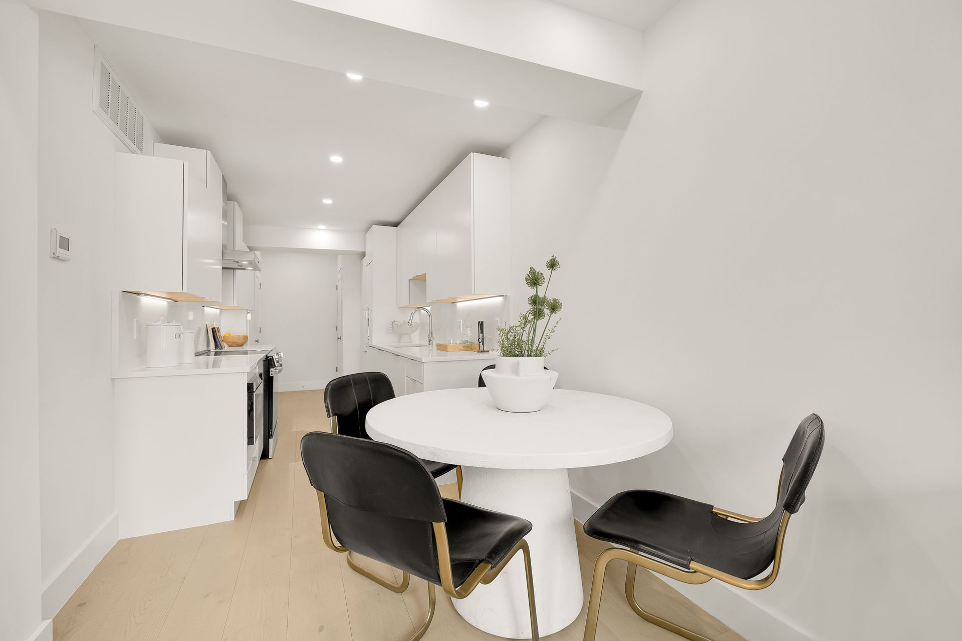 Small white kitchen with a round table and three black chairs, featuring white cabinetry and a small flower vase.