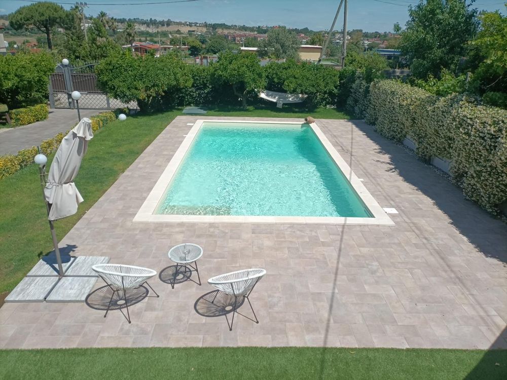 Piscina moderna nel cortile di una casa, con alberi e lettini prendisole in piena luce del sole.