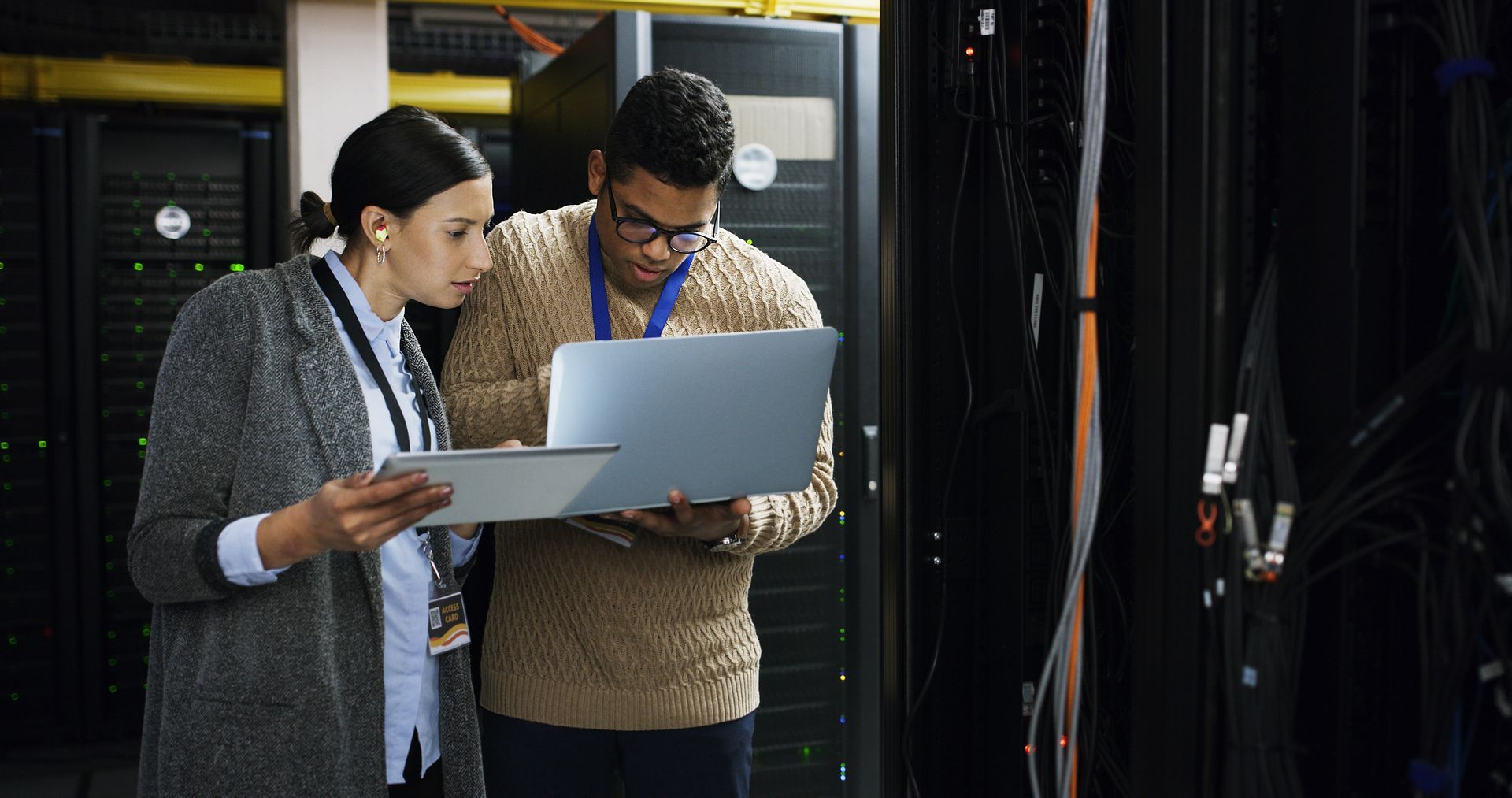 Two people in server room, examining data on laptop and tablet.