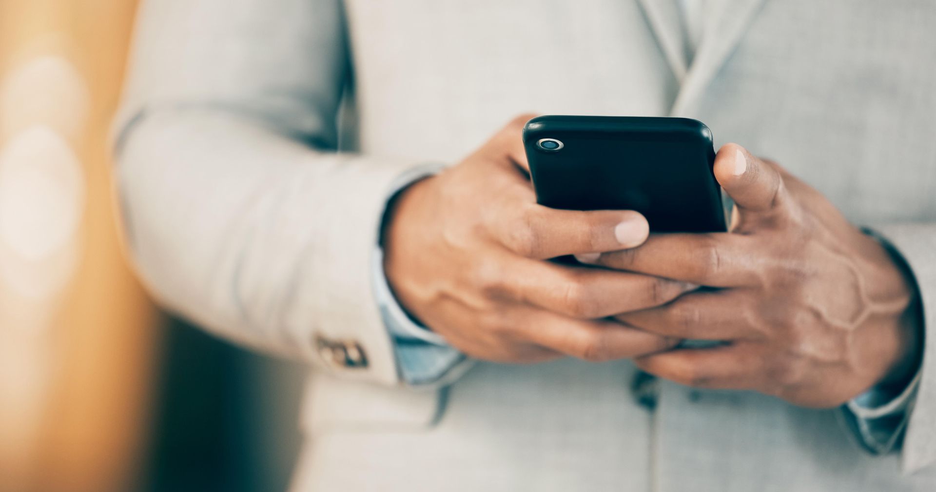 Man in light suit holds a black smartphone, typing with both hands.