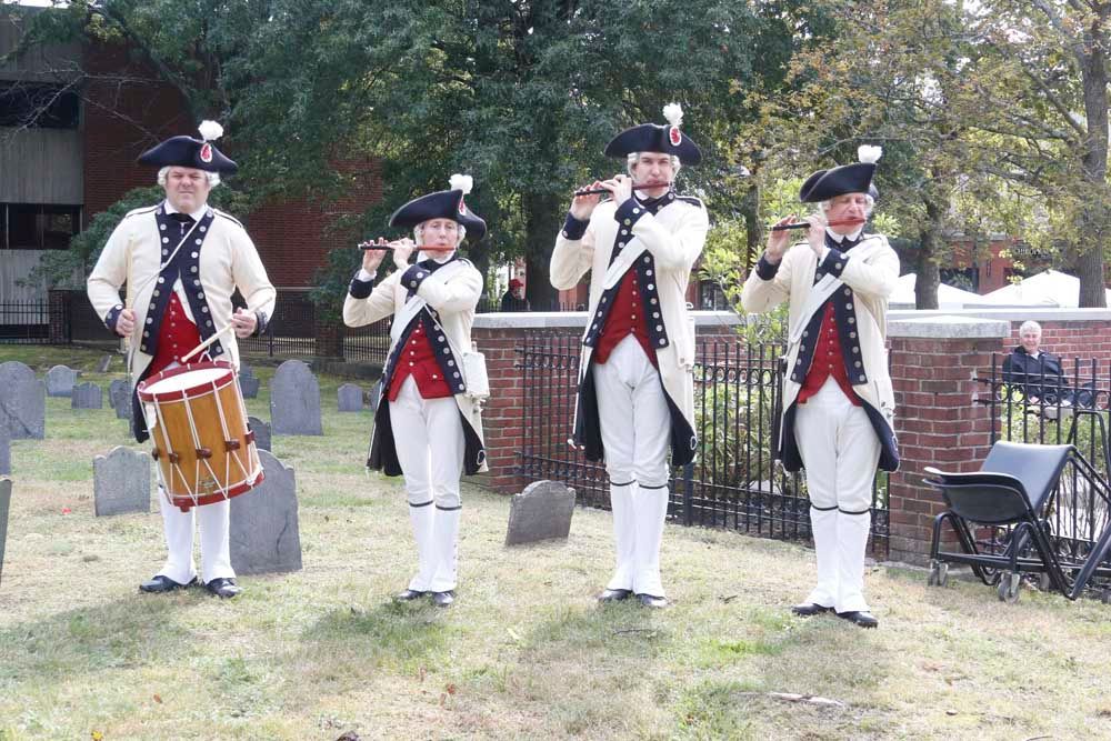 Four men in military uniforms are playing instruments in a cemetery