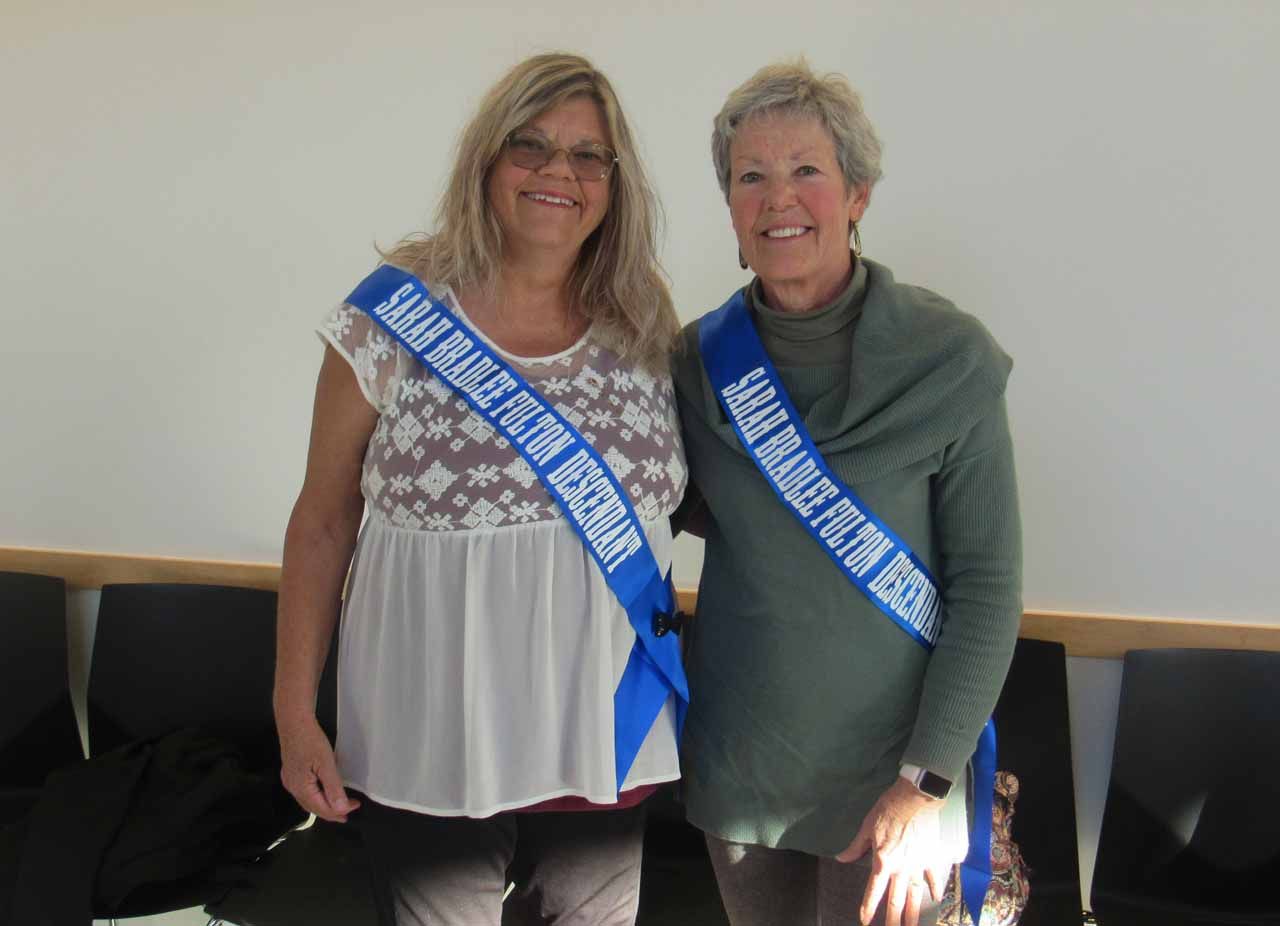 Two women standing next to each other with one wearing a sash that says james brace town resident