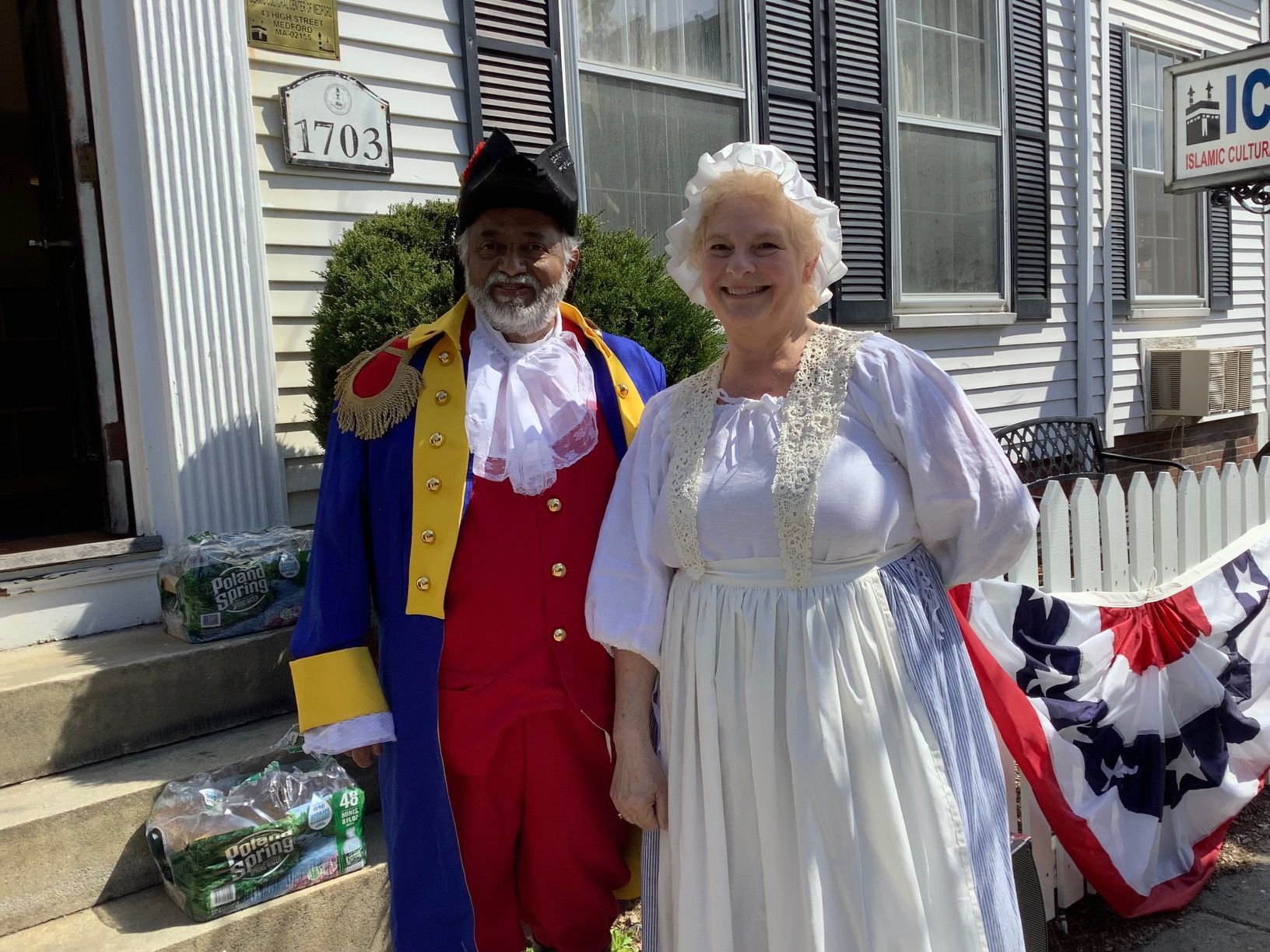 A man and a woman in historical costumes are standing in front of a house.