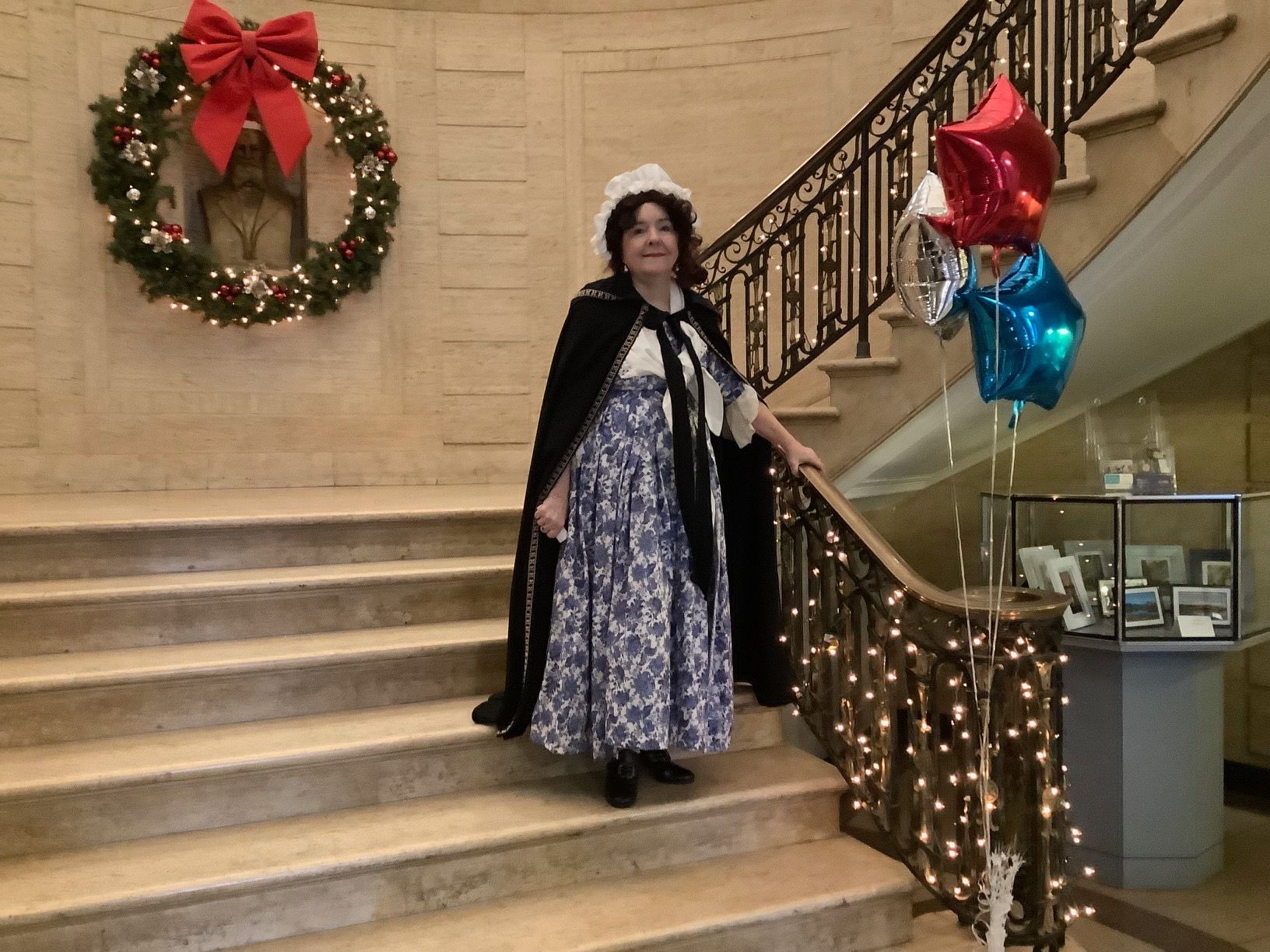 Woman in historical costume on stairs, decorated with lights and balloons, beside a wreath.