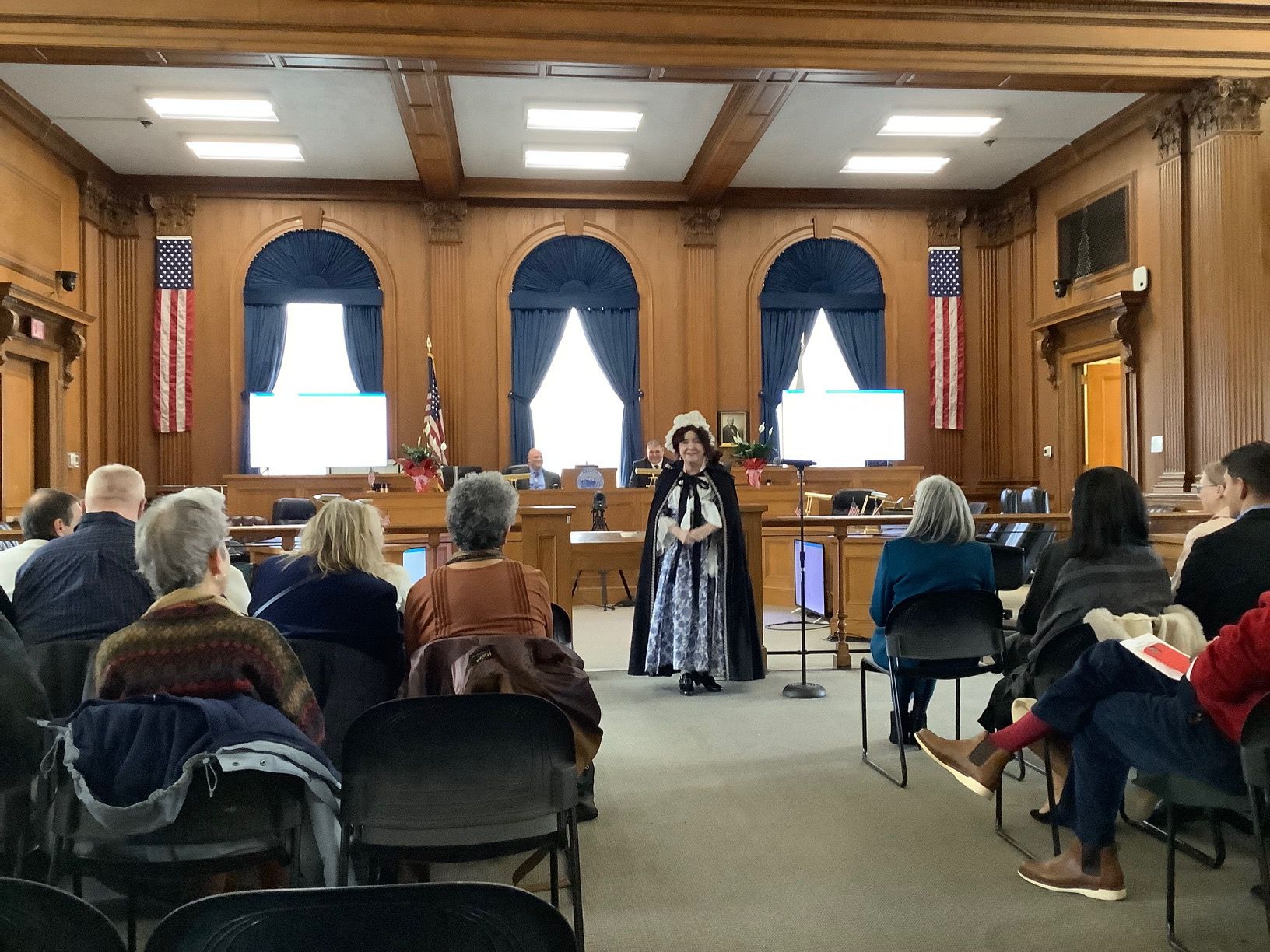 A person in costume speaking to an audience in a wood-paneled room with U.S. flags and two presentation screens.