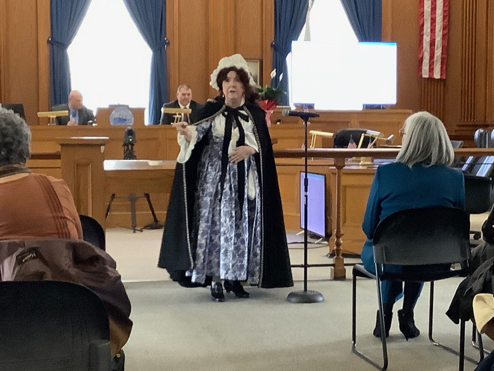 Person in historical costume speaking at podium in a courtroom. Audience members and officials in background.