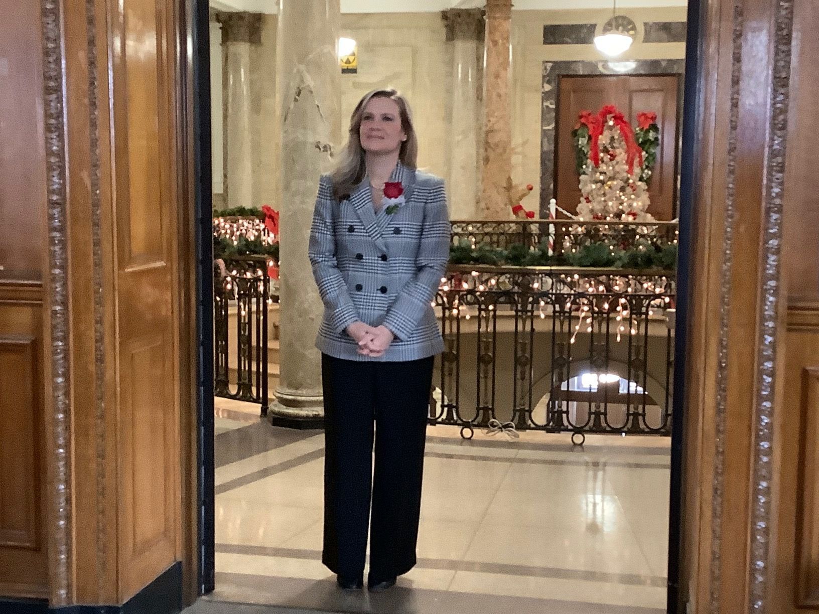 Woman in a blazer and pants stands in a doorway, hands clasped, in a festive building with holiday decorations.