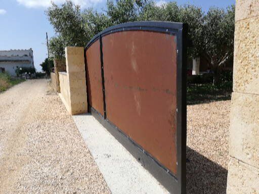 Un cancello di metallo arrugginito si trova sul ciglio di una strada sterrata, accanto a un muro di pietra.