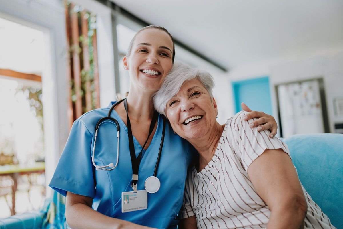 A smiling caregiver in blue scrubs with stethoscope hugs a smiling elderly woman indoors, in a home setting.