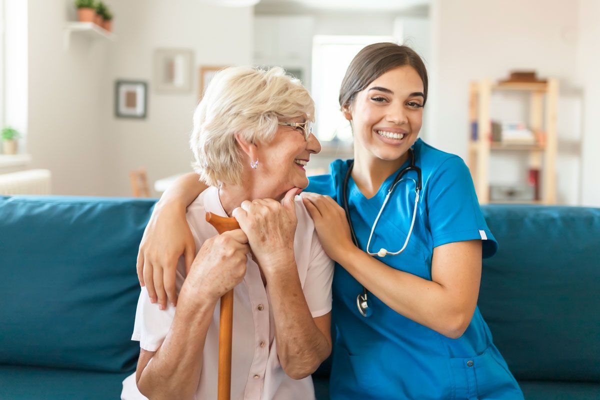 A smiling young caregiver with an arm around an older woman holding a cane on a blue couch. Both women smile.