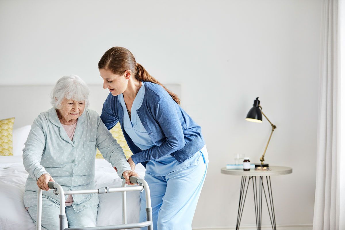 A healthcare worker assists an elderly woman using a walker in a bedroom. The worker is smiling and helping the woman stand.