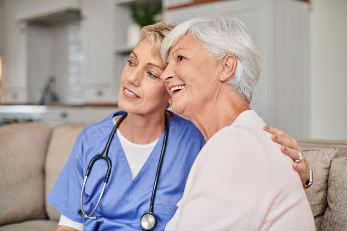 A female nurse in blue scrubs comforts an elderly woman with white hair on a couch. They are both smiling indoors.
