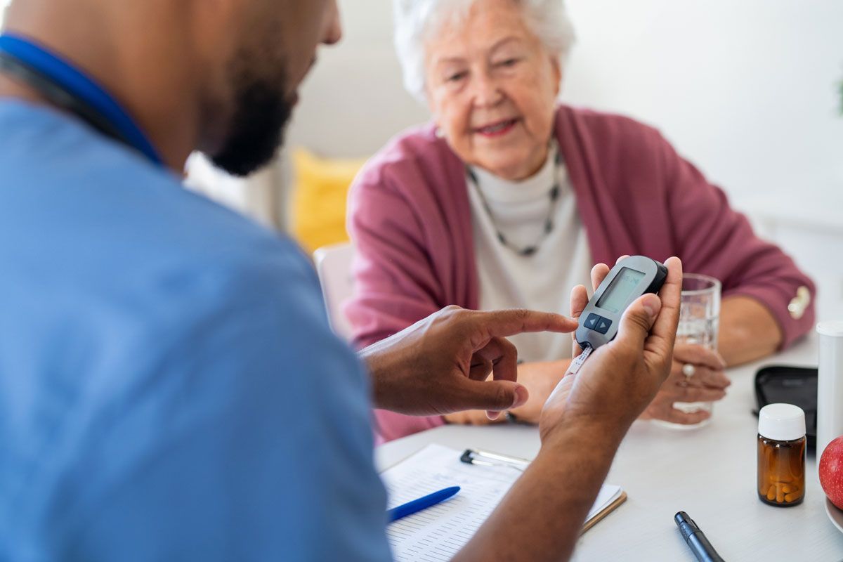 Healthcare worker showing an elderly woman a blood glucose meter. They sit at a table, the woman smiling.