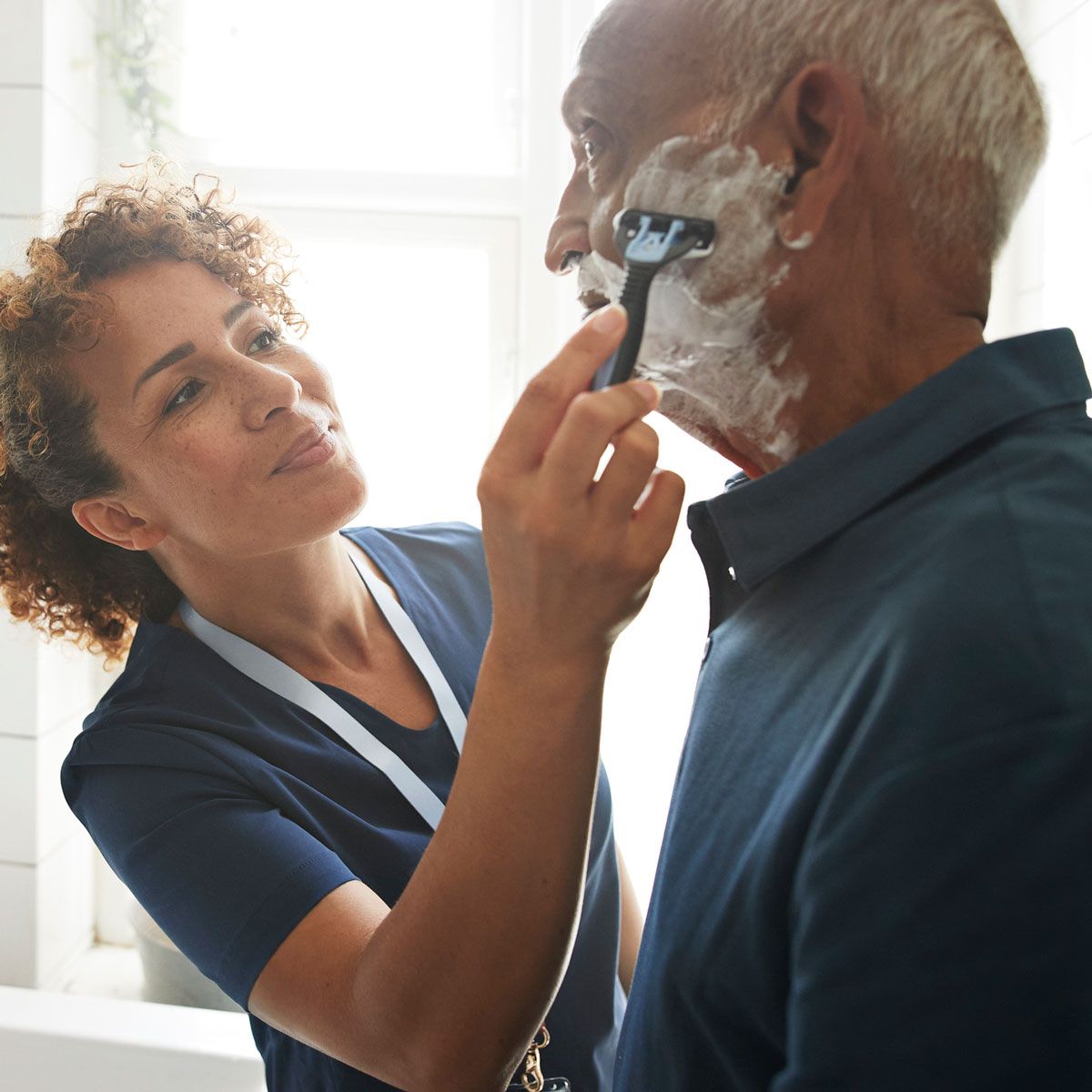 Woman assisting an elderly man to shave his face with a razor. They are in a bathroom, and the woman has a caring expression.