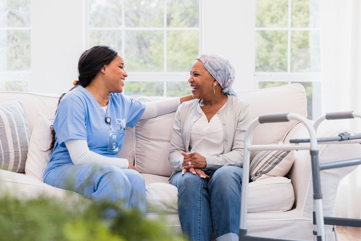 A smiling nurse in blue scrubs sits on a sofa, talking to an older woman wearing a headscarf. A walker sits beside them.