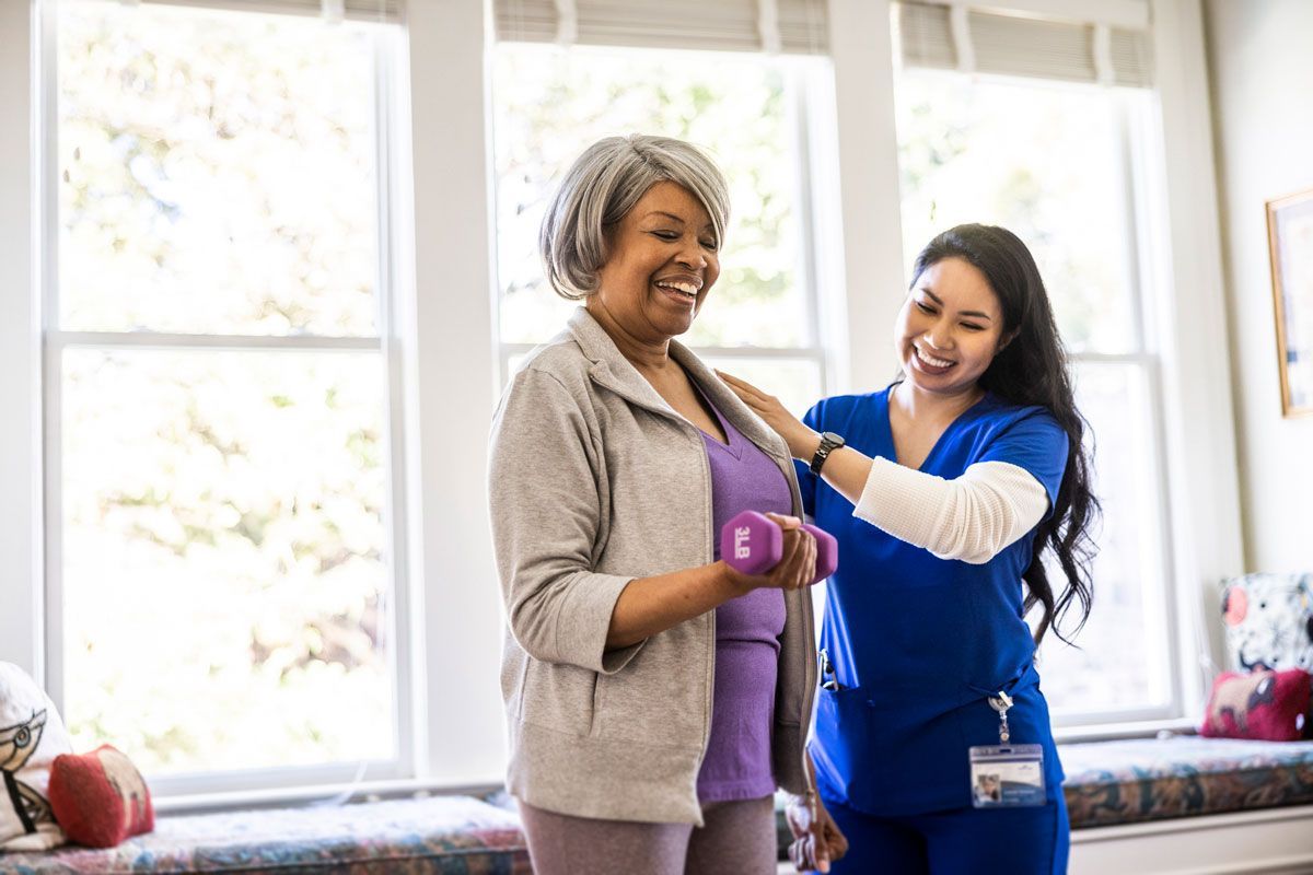 An older Black woman, smiling, lifts a purple dumbbell with guidance from a smiling Asian female caregiver in a sunlit room.