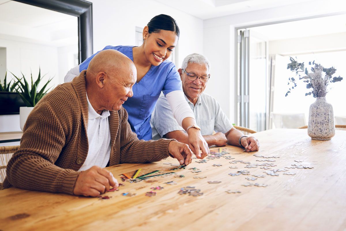 A smiling caregiver assists two older men with a jigsaw puzzle at a wooden table in a bright room.
