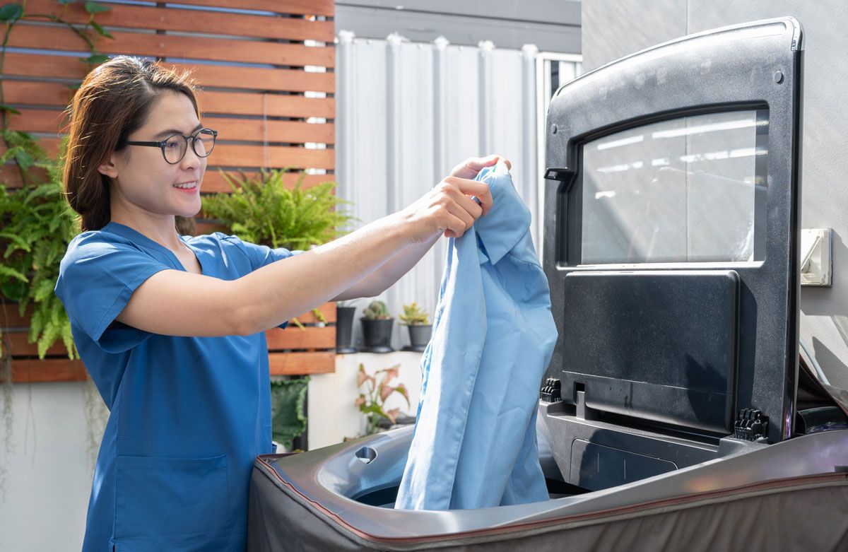 Woman in blue scrubs putting a shirt into an outdoor washing machine. She is wearing glasses and smiling in a backyard setting.