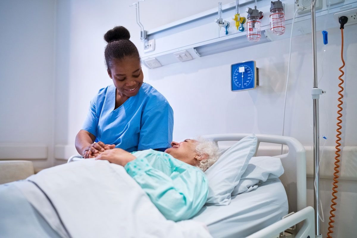 Nurse in blue scrubs smiles at an elderly patient lying in a hospital bed; both are in a bright hospital room.