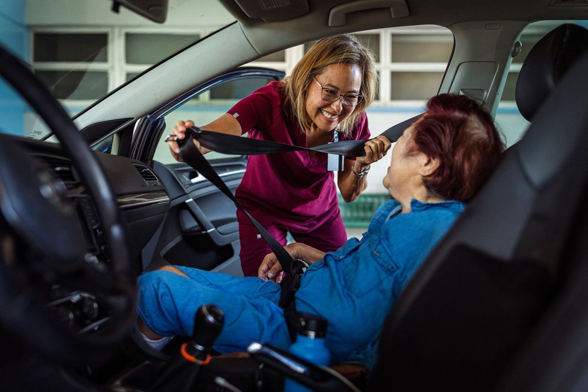 A healthcare worker fastens a seatbelt for a person in a wheelchair inside a car. Both are smiling; the worker wears pink, and the car interior is dark.